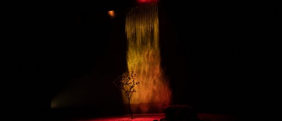 Photo, copyright Lauren Pasche, TKM : Un jeune arbre est posé sur la scène. Derrière lui, un rideau d'eau qui fait penser à une forte précipitation de pluie. Il est éclairé en orange et le sol en rouge.