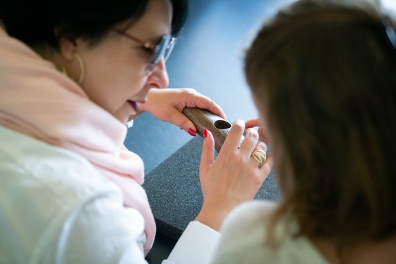 Zwei Frauen. Von einer sieht man von hinten nur den Kopf. Die andere wurde seitlich fotografiert. Von ihr sieht man die rechte Schulter und die rechte Seite des Kopfes. Trägt eine Brille. In der Hand hält sie ein Objekt, das sie ertastet. 