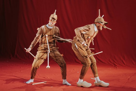 Zwei Schauspieler mit Holzhelm und langer Pinocchionase. Überall am Körper hängen unterschiedlich lange Holzstäbe.